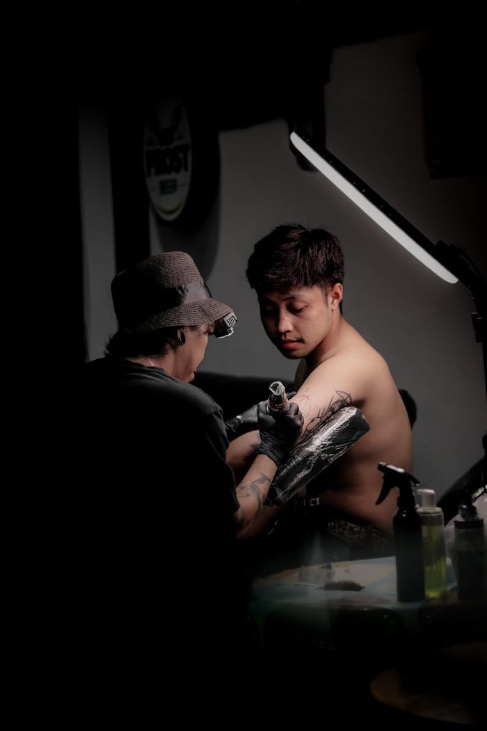 Tattoo artist applying a detailed design on a young man's arm under focused lighting.